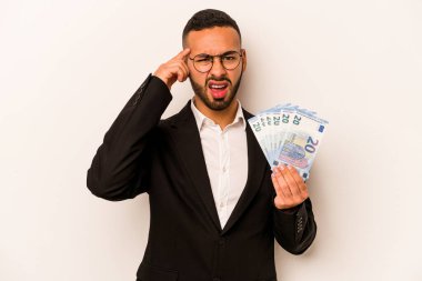 Latin kökenli genç bir adam, beyaz arka planda izole edilmiş banknotlar tutuyor ve işaret parmağıyla yaptığı hayal kırıklığı jestini gösteriyor..