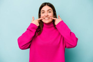 Mavi arka planda izole edilmiş genç beyaz kadın gülümsüyor, parmaklarını ağıza doğrultuyor..