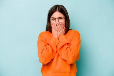 Mavi arka planda izole edilmiş genç beyaz kadın bir şeye gülüyor, elleriyle ağzını kapatıyor..