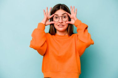 Mavi arka planda izole edilmiş genç beyaz kadın bir başarı fırsatı bulmak için gözlerini açık tutuyor..