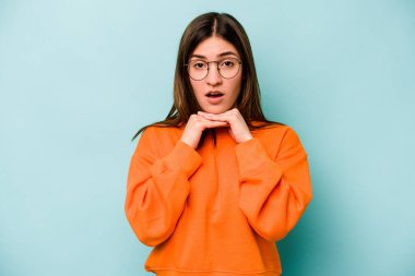 Kafkasyalı genç bir kadın mavi arka planda izole edilmiş şans için dua ediyor, şaşırmış ve ağzını açıp önüne bakıyor..