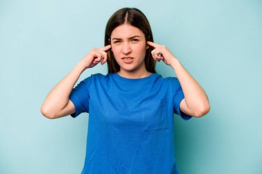 Genç beyaz kadın mavi arka planda izole edilmiş parmaklarıyla kulaklarını kapatıyor, gürültülü bir ortam yüzünden stresli ve çaresiz..