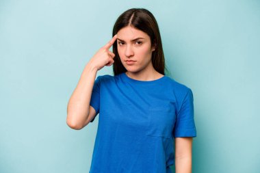 Mavi arka planda izole edilmiş genç beyaz kadın şakağı parmağıyla işaret ediyor, düşünüyor ve bir göreve odaklanıyor..