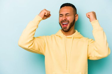Mavi arka planda izole edilmiş genç İspanyol bir adam zaferden sonra yumruk kaldırıyor, kazanan konsept..