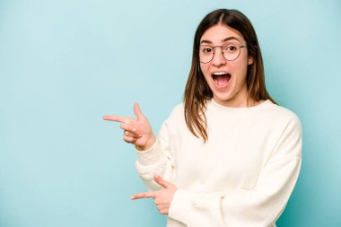 Mavi arka planda izole edilmiş genç beyaz kadın işaret parmağıyla işaret ederek heyecanlanıyor..