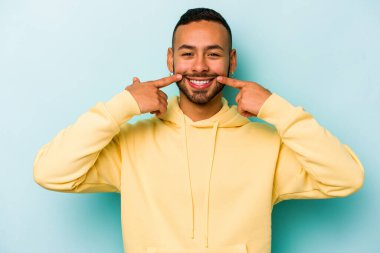 Mavi arka planda izole edilmiş genç İspanyol adam gülümsüyor, parmaklarını ağıza doğrultuyor..