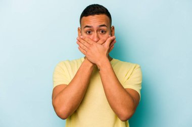 Mavi arka planda izole edilmiş genç İspanyol adam elleriyle ağzını kapatırken şok oldu..