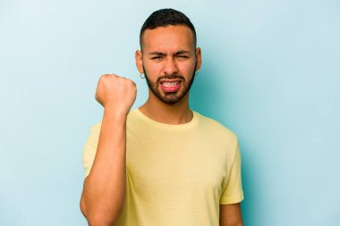 Mavi arka planda izole edilmiş genç İspanyol adam kameraya yumruk gösteriyor, agresif yüz ifadesi..