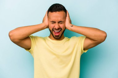 Genç İspanyol bir adam mavi arka planda izole edilmiş kulaklarını elleriyle kapatıyor çok fazla ses duymamaya çalışıyor..