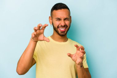 Mavi arka planda izole edilmiş genç İspanyol bir adam pençelerini bir kediyi taklit ederken gösteriyor, agresif bir hareket..