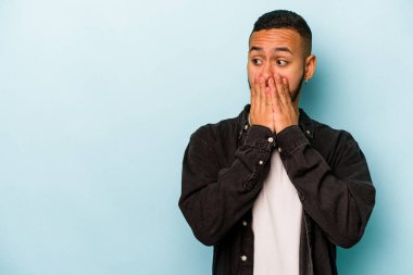 Genç İspanyol adam mavi arka planda izole edilmiş. Düşünceli bir şekilde bir fotokopi odasına bakıyor. Ağzını eliyle kapatıyor..