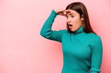 Pembe arka planda izole edilmiş genç beyaz kadın uzaklara bakıyor ve elini alnına koyuyor..