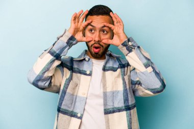 Genç İspanyol bir adam mavi arka planda izole edilmiş. Başarı fırsatı bulmak için gözlerini açık tutuyor..