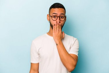 Mavi arka planda izole edilmiş genç İspanyol adam şok olmuş, ağzı ellerle kaplanmış, yeni bir şey keşfetmek için sabırsızlanıyor..