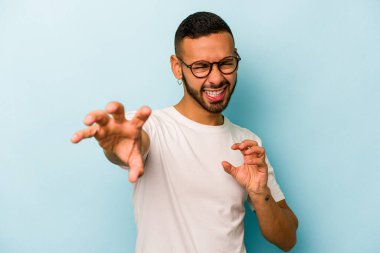 Mavi arka planda izole edilmiş genç İspanyol bir adam pençelerini bir kediyi taklit ederken gösteriyor, agresif bir hareket..