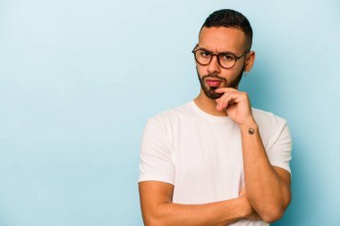 Genç İspanyol adam mavi arka planda izole edilmiş şüpheli ve şüpheci bir ifadeyle yana bakıyor..