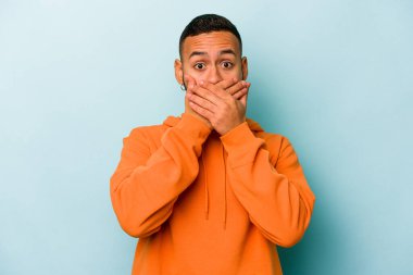 Mavi arka planda izole edilmiş genç İspanyol adam elleriyle ağzını kapatırken şok oldu..