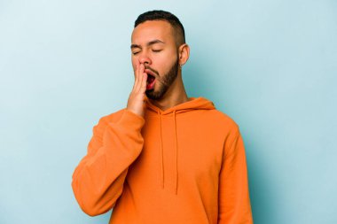 Mavi arka planda izole edilmiş genç İspanyol adam esniyor. Ağzı eliyle kapatan yorgun bir hareket sergiliyor..