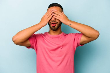 Mavi arka planda izole edilmiş genç İspanyol adam elleriyle gözlerini kapatmaktan korkuyor..