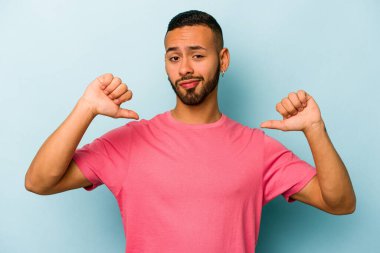 Mavi arka planda izole edilmiş genç İspanyol adam baş parmağını aşağı gösteriyor, hayal kırıklığı konsepti.