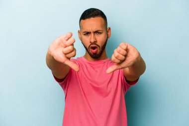 Mavi arka planda izole edilmiş genç İspanyol adam baş parmağını aşağı gösteriyor ve hoşlanmadığını ifade ediyor..