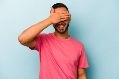 Mavi arka planda izole edilmiş genç İspanyol adam elleriyle gözlerini kapatıyor, geniş bir gülümsemeyle bir sürpriz bekliyor..