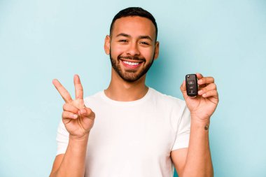Mavi arka planda izole edilmiş araba anahtarlarını tutan genç İspanyol adam neşeli ve kaygısız parmaklarıyla barış sembolü gösteriyor..