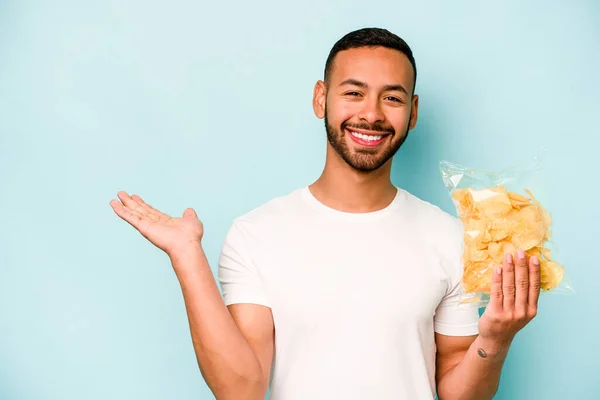 Young hispanic man holding a bag of chips isolated on blue background showing a copy space on a palm and holding another hand on waist.