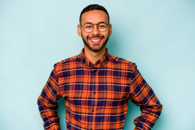 Latin kökenli genç adam mavi arka planda izole edilmiş. Kendine güveniyor. Kalçaları tutuyor..