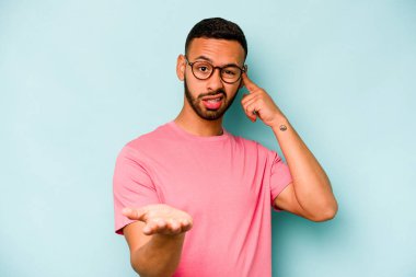 Genç İspanyol bir adam mavi arka planda izole edilmiş ve elinde bir ürün tutuyor..