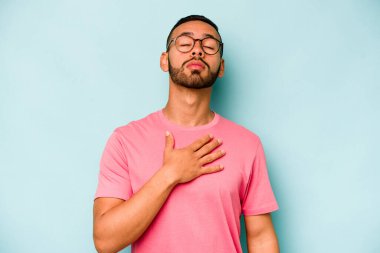 Genç İspanyol bir adam mavi arka planda izole edilmiş yemin ediyor, elini göğsüne koyuyor..