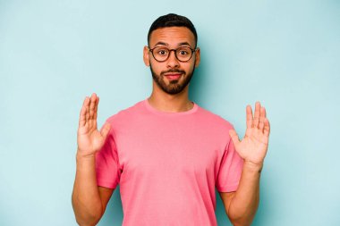 Mavi arka planda izole edilmiş genç İspanyol adam işaret parmaklarıyla küçük bir şey tutuyor, gülümsüyor ve kendinden emin..