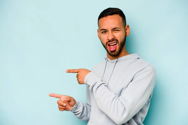 Young hispanic man isolated on blue background pointing with forefingers to a copy space, expressing excitement and desire.