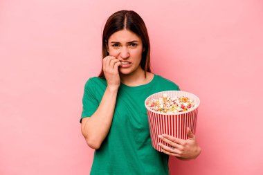 Elinde patlamış mısır tutan beyaz bir kadın pembe arka planda izole edilmiş tırnak yiyor, gergin ve endişeli..