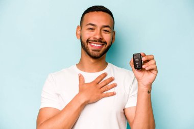 Mavi arka planda izole edilmiş araba anahtarlarını tutan İspanyol genç adam yüksek sesle gülüyor ve elini göğsüne koyuyor..