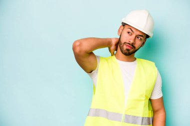 Latin kökenli genç işçi mavi arka planda izole edilmiş kafasının arkasına dokunuyor, düşünüyor ve bir seçim yapıyor..
