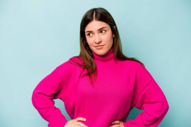 Kafkasyalı genç bir kadın mavi arka planda izole edilmiş, kafası karışmış, şüpheli ve kendinden emin değil..