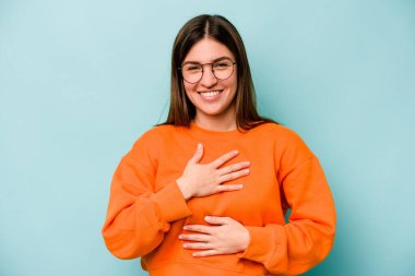 Mavi arka planda izole edilmiş genç beyaz kadın mutlu bir şekilde gülüyor ve ellerini karnının üstünde tutarak eğleniyor..