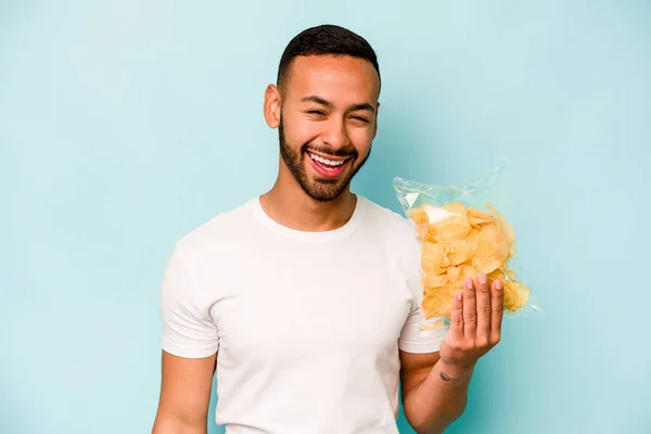 Young hispanic man holding a bag of chips isolated on blue background laughing and having fun.