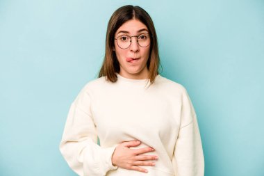 Mavi arka planda izole edilmiş genç beyaz kadın karnına dokunuyor, nazikçe gülümsüyor, yemek yiyor ve tatmin oluyor..