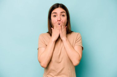 Mavi arka planda izole edilmiş genç beyaz kadın şok olmuş, ağzı ellerle kaplanmış, yeni bir şey keşfetmeye hevesli..