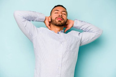 Genç İspanyol adam mavi arka planda izole edilmiş. Kendine güveniyor, elleri başının arkasında..