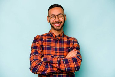 Mavi arka planda kendini güvende hisseden, kararlılıkla kollarını kavuşturan genç İspanyol bir adam..