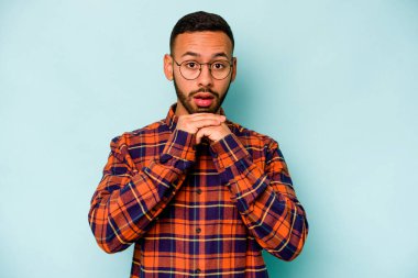 Latin kökenli genç adam mavi arka planda izole edilmiş şans için dua ediyor, şaşırmış ve ağzı açık bir şekilde önüne bakıyor..