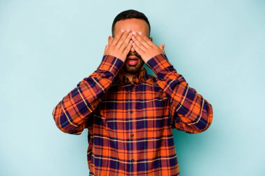 Mavi arka planda izole edilmiş genç İspanyol adam elleriyle gözlerini kapatmaktan korkuyor..