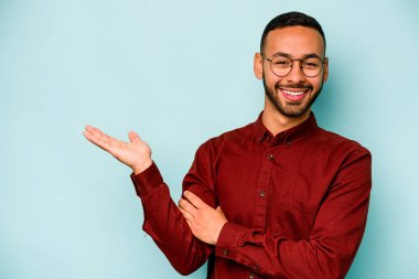 Mavi arka planda izole edilmiş genç İspanyol bir adam avucunun içinde bir fotokopi alanı ve belinde bir el daha gösteriyor..
