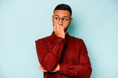 Mavi arka planda izole edilmiş, tırnak yiyen, gergin ve endişeli genç bir adam..