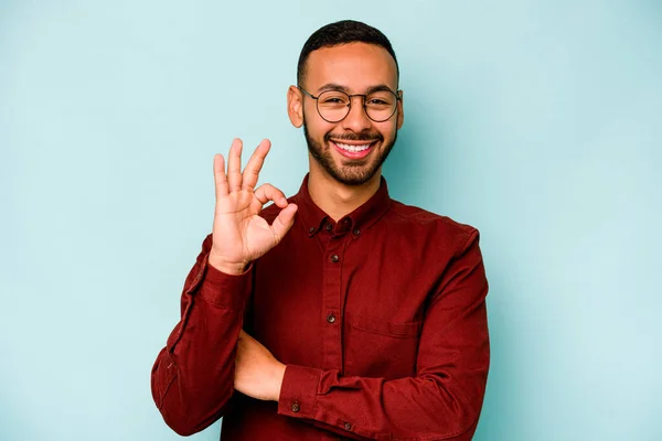 Young hispanic man isolated on blue background winks an eye and holds an okay gesture with hand.