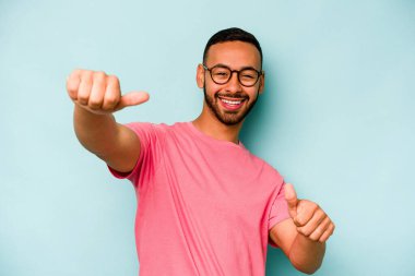 Genç İspanyol adam mavi arka planda izole edilmiş. Baş parmağını kaldırıyor, gülümsüyor ve kendinden emin..