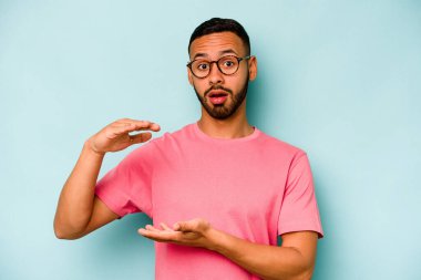 Mavi arka planda izole edilmiş genç İspanyol adam iki eliyle bir şey tutuyor, ürün sunumu..